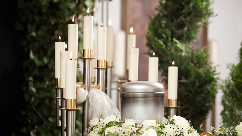 Silberne Urne umgeben von weißen Rosen und Kerzen auf einem Tisch, im Hintergrund grüne Pflanzen.