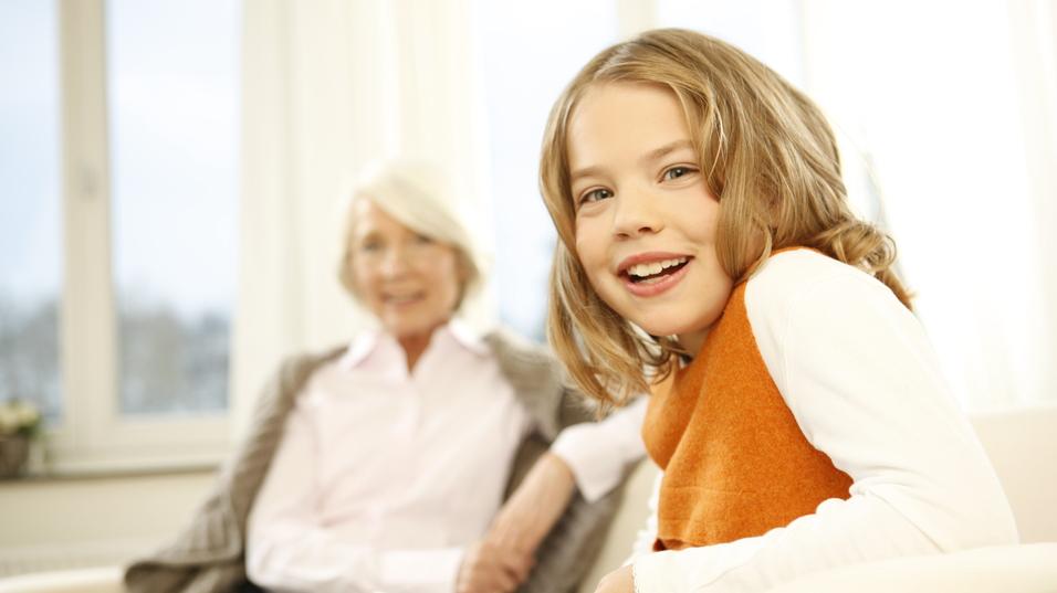 Lächelndes Mädchen mit Buch, im Hintergrund eine ältere Frau, beide in heller Wohnumgebung.